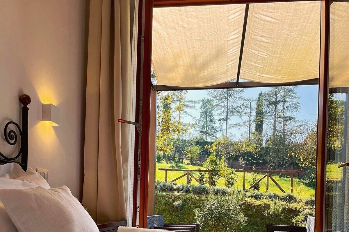 Hotellihuone, jossa on sänky, valkoiset pyyhkeet, yöpöytä ja suuri ikkuna, josta avautuu näkymä puutarhaan.