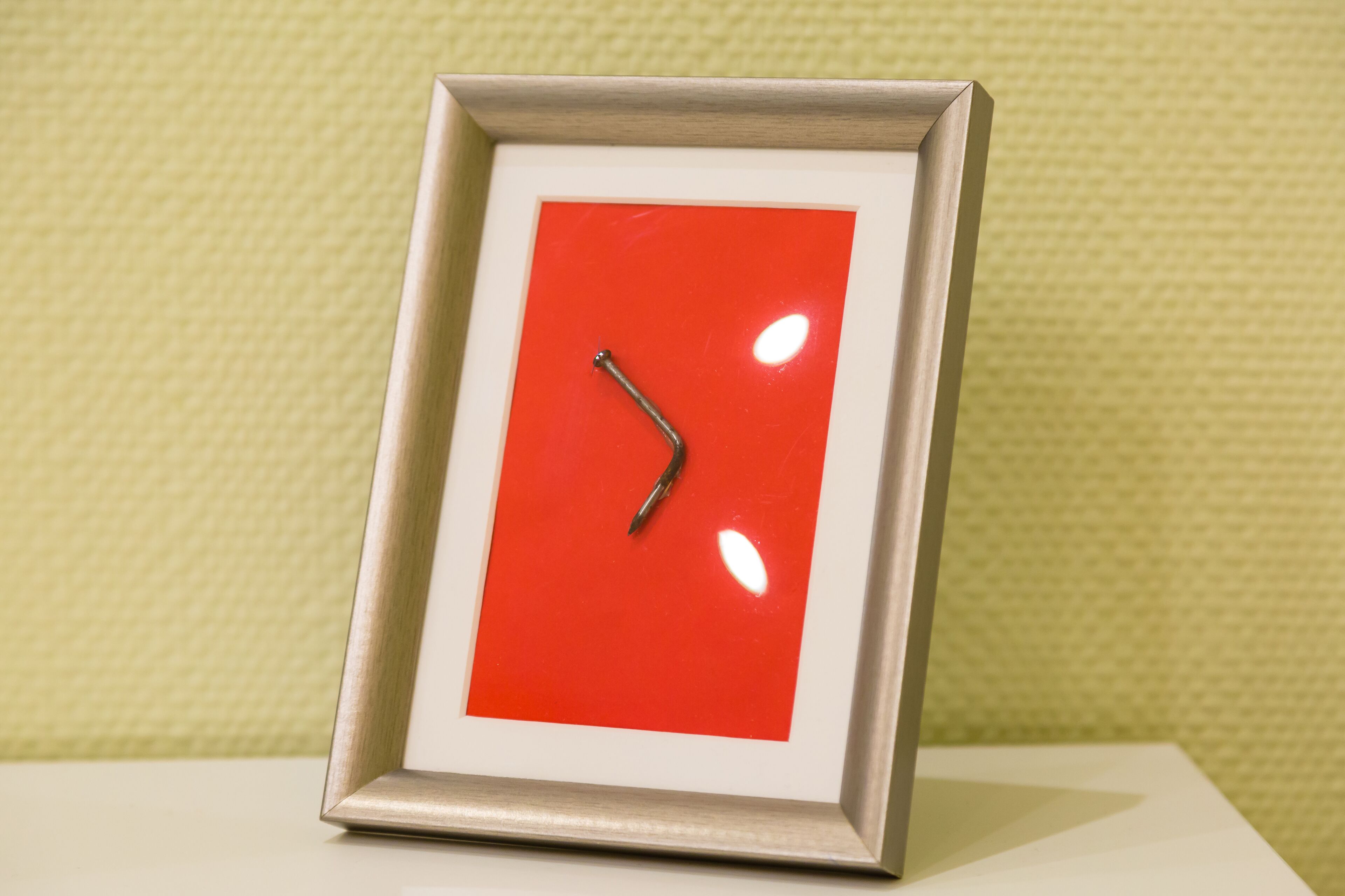 Un reloj en marco con esfera roja y manecilla metálica, sobre una pared con textura.