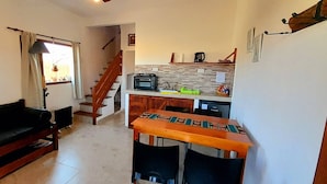 Un salon comprenant un canapé, une table en bois et un escalier menant à un coin cuisine équipé d’une radio et d’une bouilloire.
