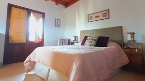 Une chambre avec un plafond en bois, un lit recouvert d’une couverture rose, une table de chevet avec une lampe et une fenêtre avec des rideaux.