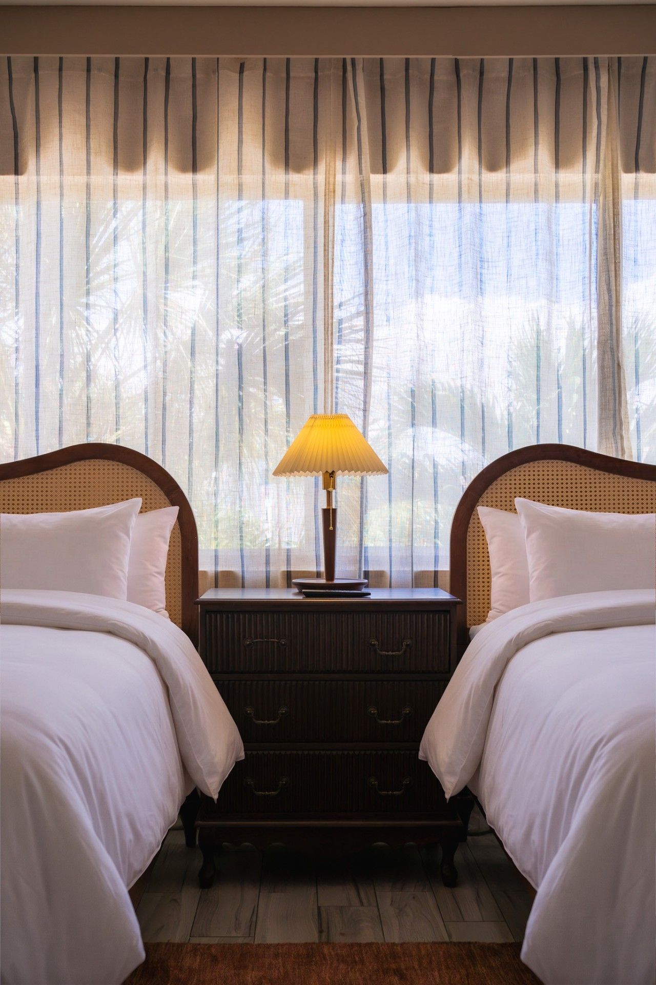 A hotel room with two beds, a nightstand with a lamp, and a window with curtains.