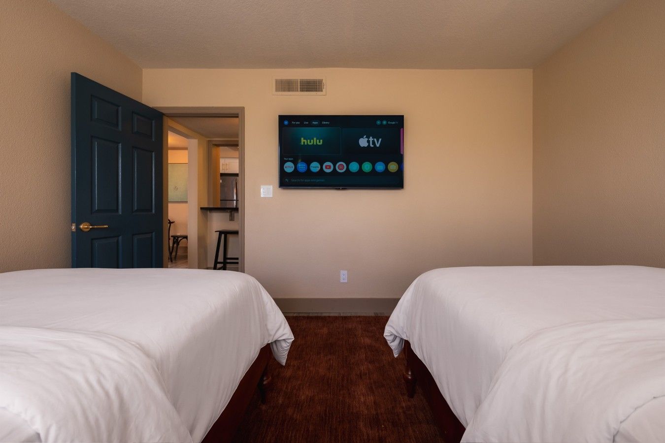 A hotel room with two beds, a TV mounted on the wall displaying Hulu and Apple TV, and a door leading to a kitchenette area.