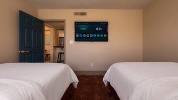 A hotel room with two beds, a TV mounted on the wall displaying Hulu and Apple TV, and a door leading to a kitchenette area.