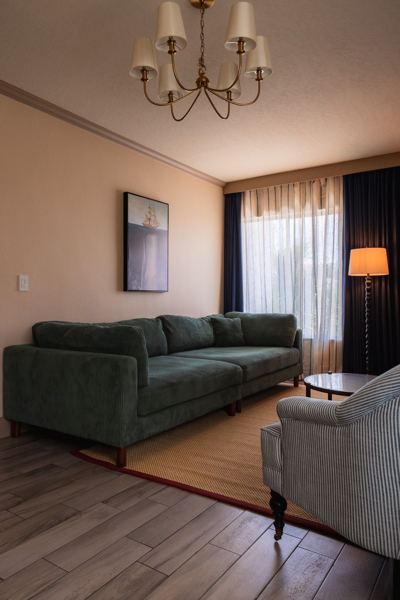 Una sala de estar con un sofá verde, un sillón a rayas, una mesa de centro redonda, una araña, un cuadro y una ventana con cortinas.