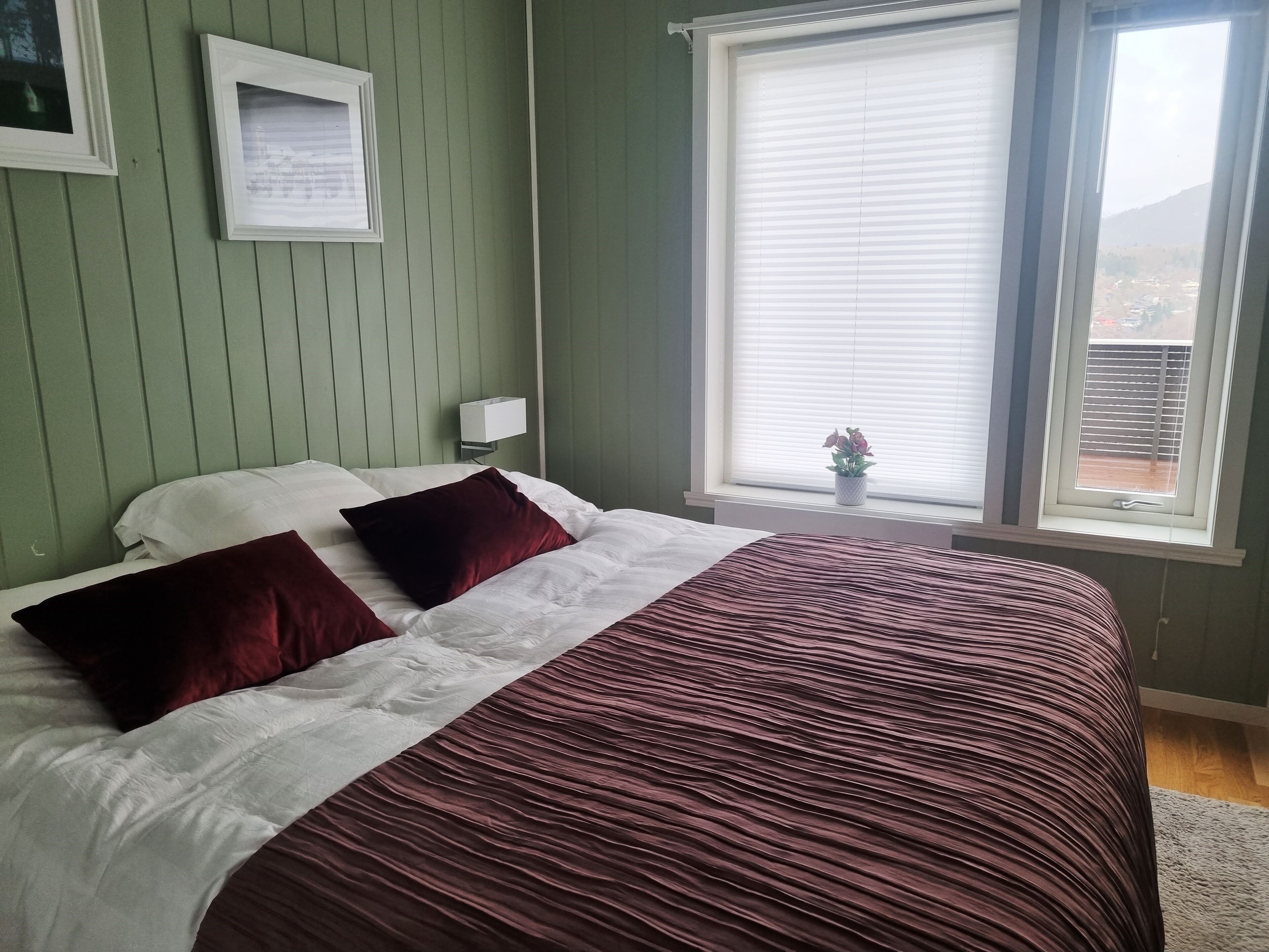 Un dormitorio con cama, paredes verdes, ventana con persianas, mesita de noche y un cuadro enmarcado en la pared.