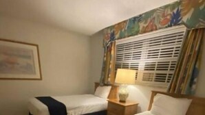 A hotel room with two beds, a nightstand, a lamp, and a window with blinds. - Turtle Reef Club Resort B (Jensen Beach)