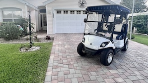 En golfbil parkerad framför ett hus med ett garage, en välskött gräsmatta och en liten trädgård med en fågelmatare.