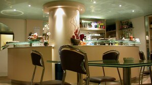 A modern bar area with a glass table and chairs, a decorative pillar, and shelves with bottles and glasses. - Hotel Concordia (Loano)
