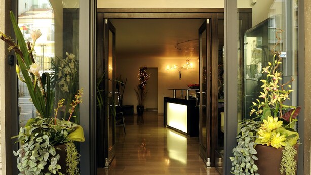 A modern hotel lobby with a wooden floor, a reception desk, and potted plants on either side of the entrance. - Hotel Concordia (Loano)