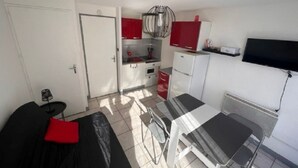 Quarto compacto com cama, uma mesinha, cadeira, cozinha compacta e televisão fixada na parede.