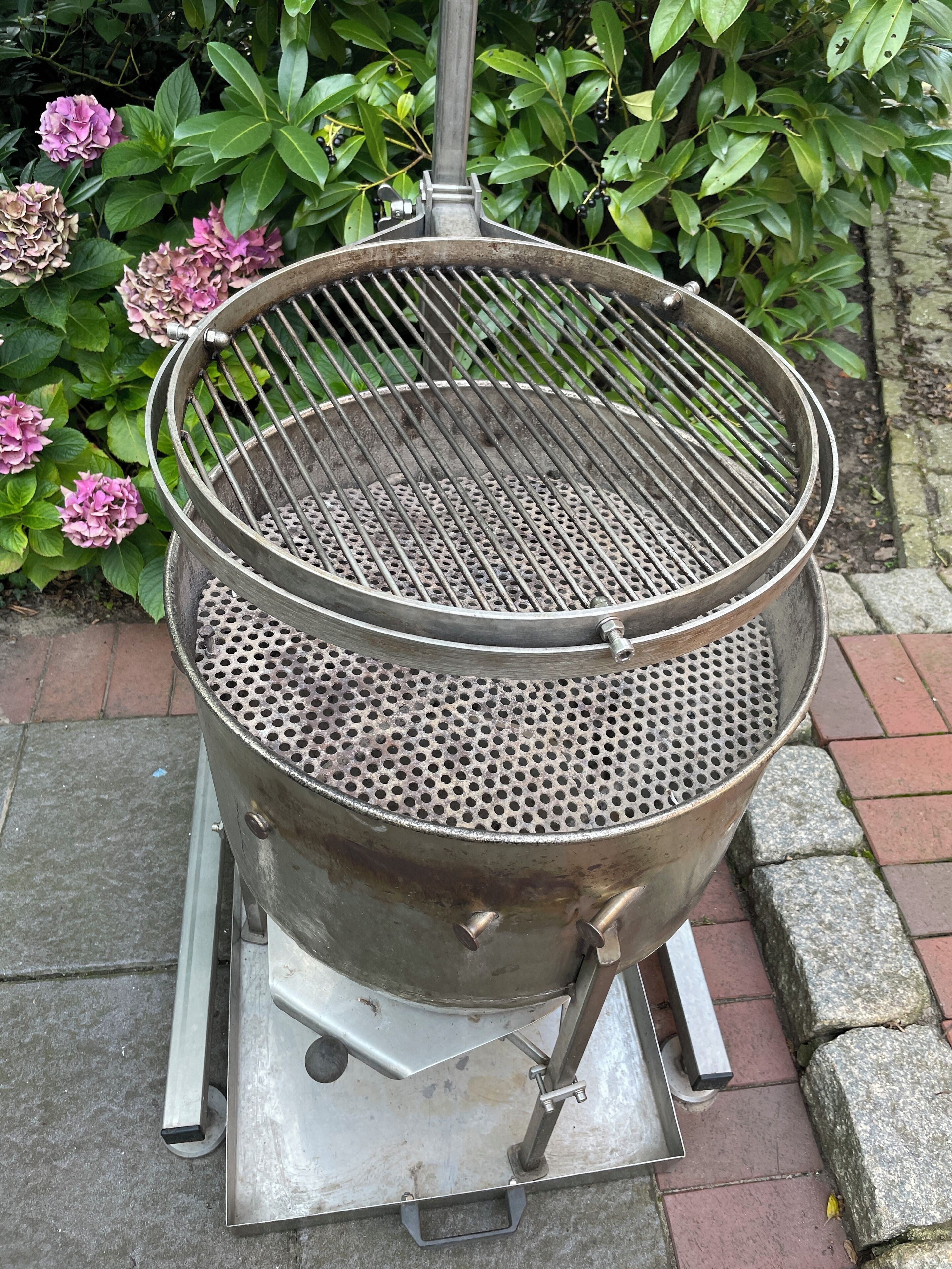 A portable grill with a perforated grate, set on a metal stand with wheels, positioned outdoors with a brick pathway and greenery in the background.