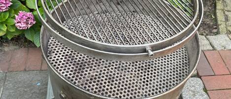 A portable grill with a perforated grate, set on a metal stand with wheels, positioned outdoors with a brick pathway and greenery in the background.