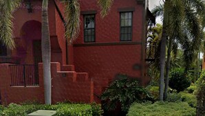 Un edificio rojo con un camino de ladrillo que conduce a la entrada, rodeado de palmeras y follaje.