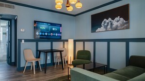 Una habitación moderna con televisión de pantalla plana mostrando aplicaciones de transmisión, un sofá verde, una mesita pequeña, dos sillas y un cuadro de nubes en la pared.