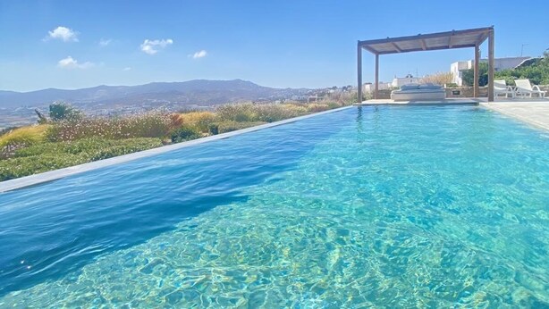 A clear blue pool with a pergola and lounge chairs, surrounded by a scenic landscape. - Aura villa A story to tell  (Mykonos)