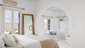 A bedroom with a large bed, a sitting area with a chair, and a view of the countryside through the window. - Aura villa A story to tell  (Mykonos)
