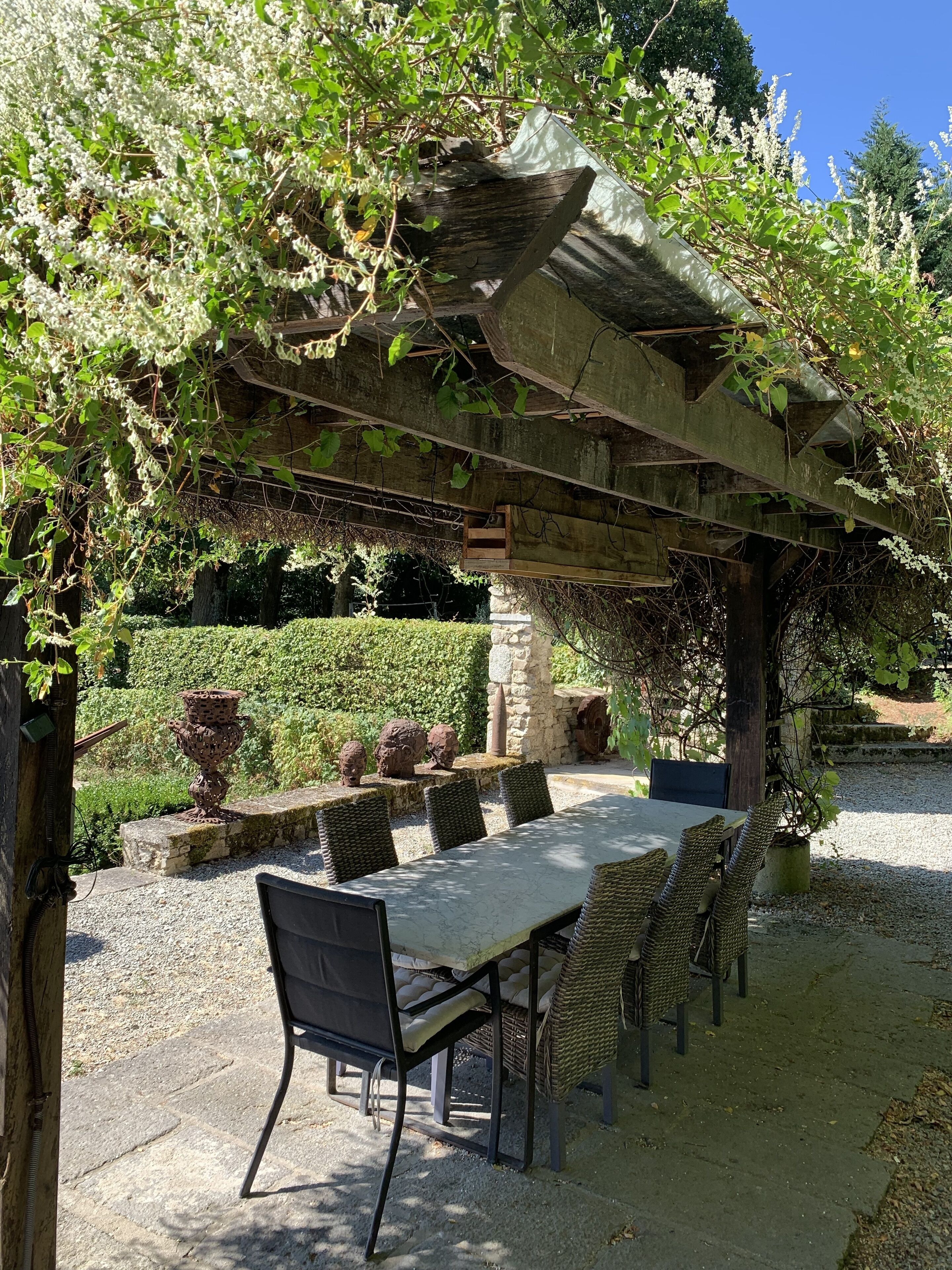 Un patio exterior con una pérgola, columnas de piedra y una mesa con sillas de mimbre.