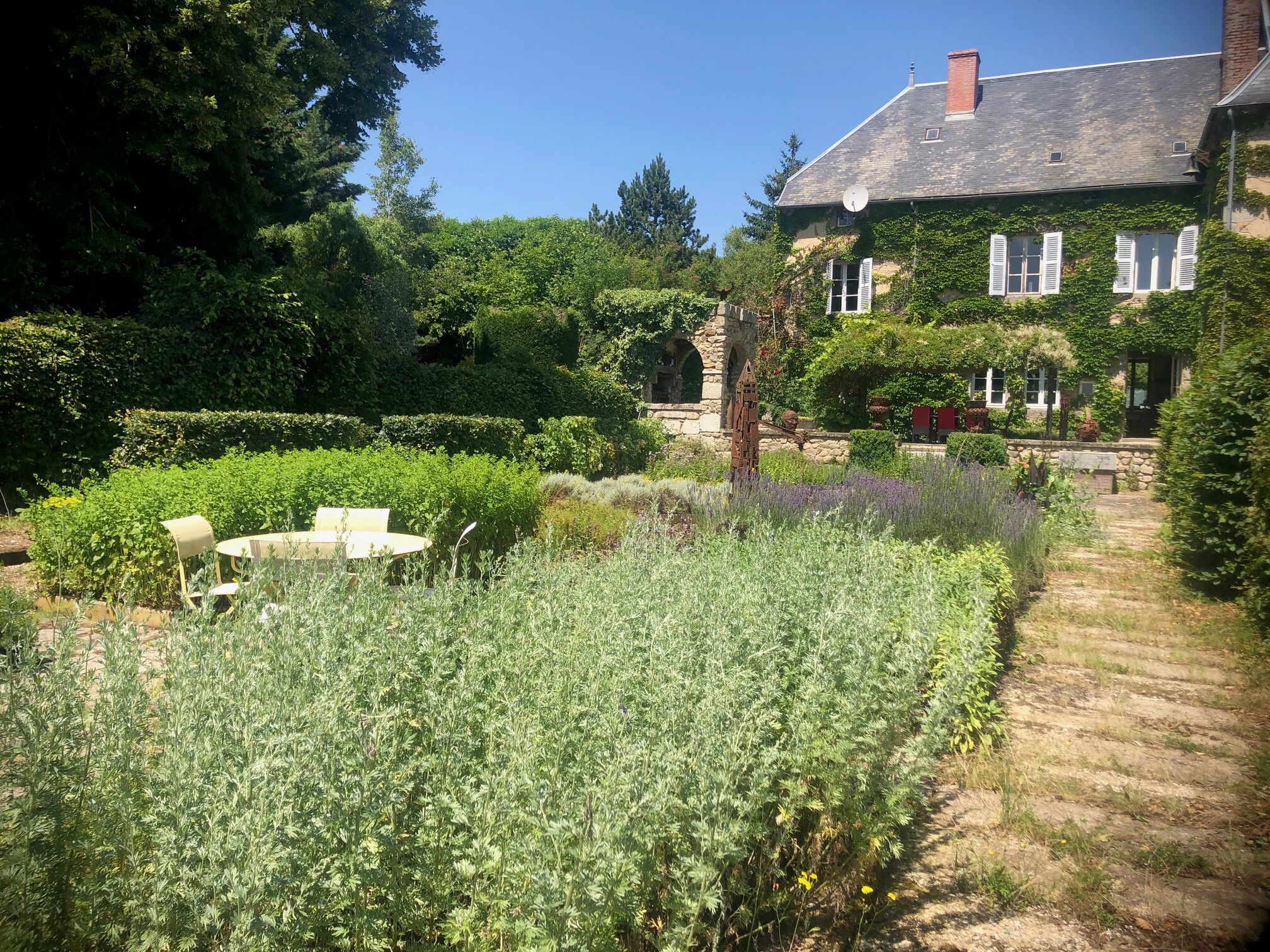 A garden with a table and chairs, a stone pathway, and a building with a sloped roof and shutters.