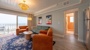 Een zithoek met een bank, twee fauteuils, een salontafel en een kroonluchter. Er hangen twee schilderijen aan de muur, een balkon met een hekwerk en uitzicht op de oceaan.