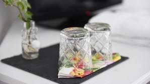 Dos saleros y pimenteros de vidrio sobre un posavasos negro con un tapete colorido debajo, colocados sobre una superficie blanca con fondo desenfocado.