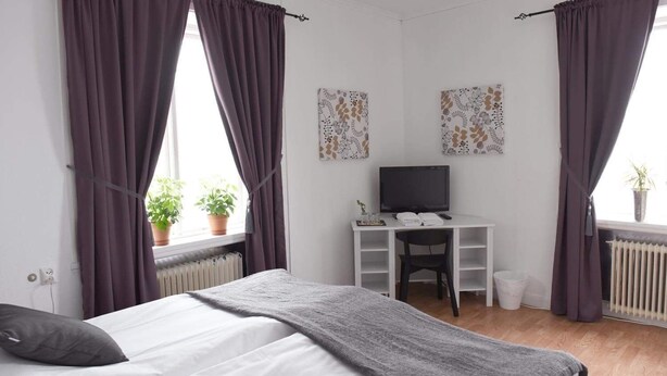 Un dormitorio con cama, escritorio, televisor y dos ventanas con cortinas.