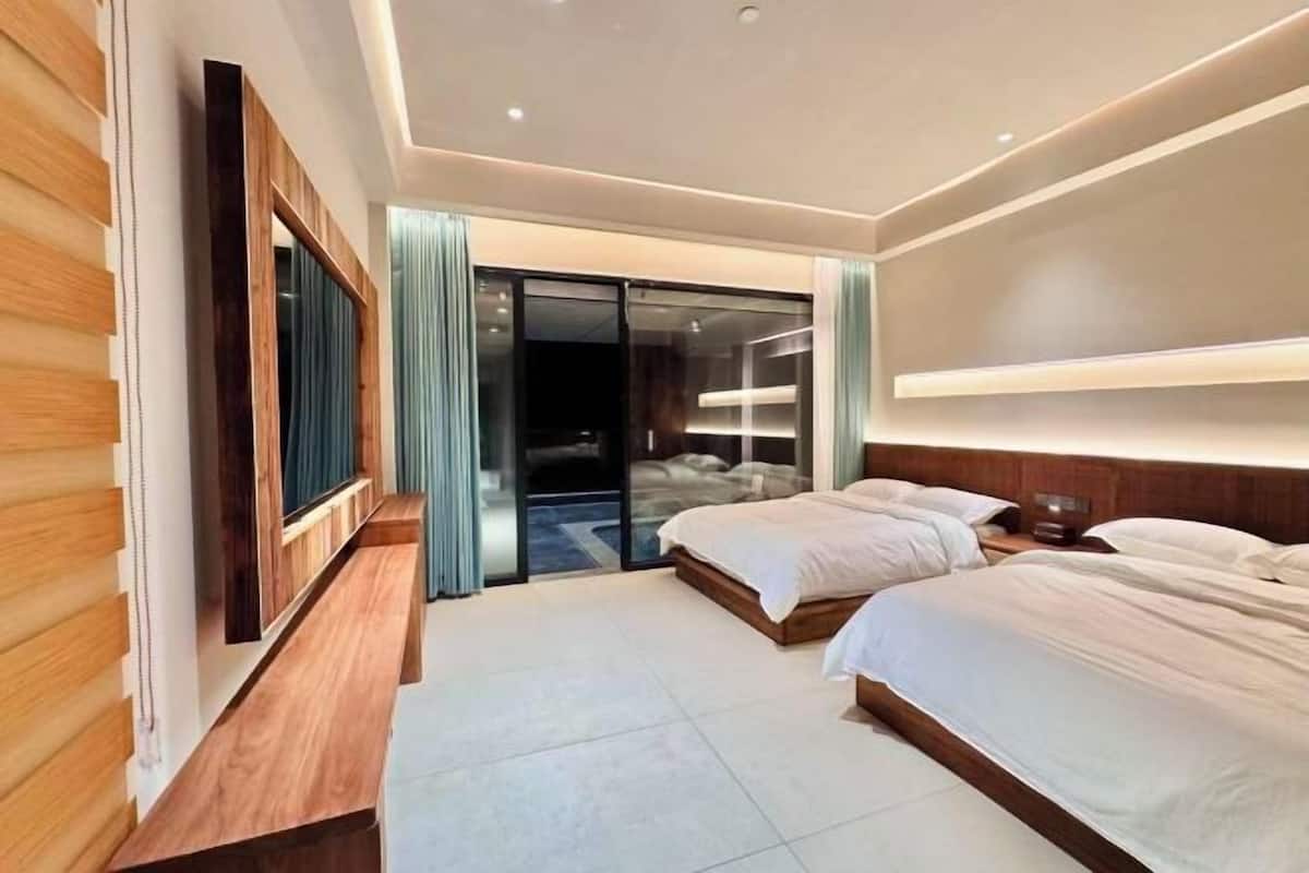A modern hotel room with a wooden bench, two beds, a large sliding glass door, and a ceiling light fixture.