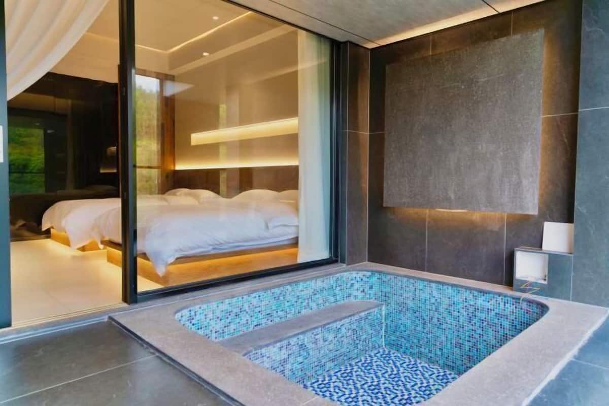 A modern bathroom with a glass-enclosed shower, a large bathtub with mosaic tiles, and a visible bedroom with a bed and curtains.