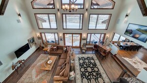 A spacious living area with a large sofa, a dining table, and a chandelier. There are multiple windows providing a view of the outdoors. - Madison River House - Ennis, MT (Madison County)