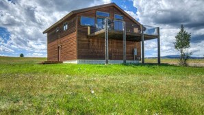A wooden cabin with a balcony, surrounded by a grassy field and a cloudy sky. - 2-bedroom cabin with views of the Madison River! (Madison County)