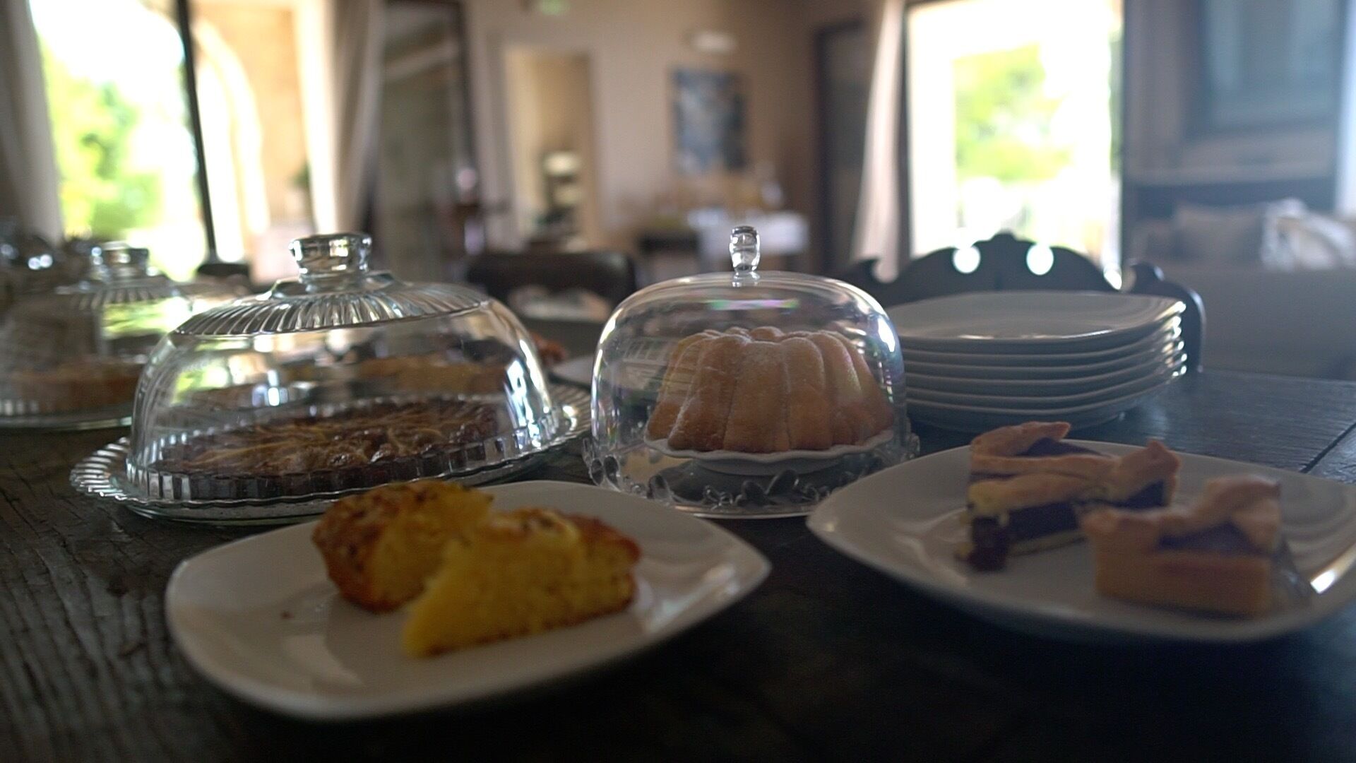 A table with various desserts under glass domes, plates with pastries, and a stack of plates in the background.
