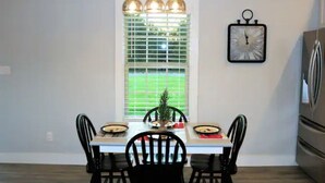 Un coin repas comprenant une table, des chaises, des luminaires suspendus, une fenêtre avec des stores et une horloge au mur.