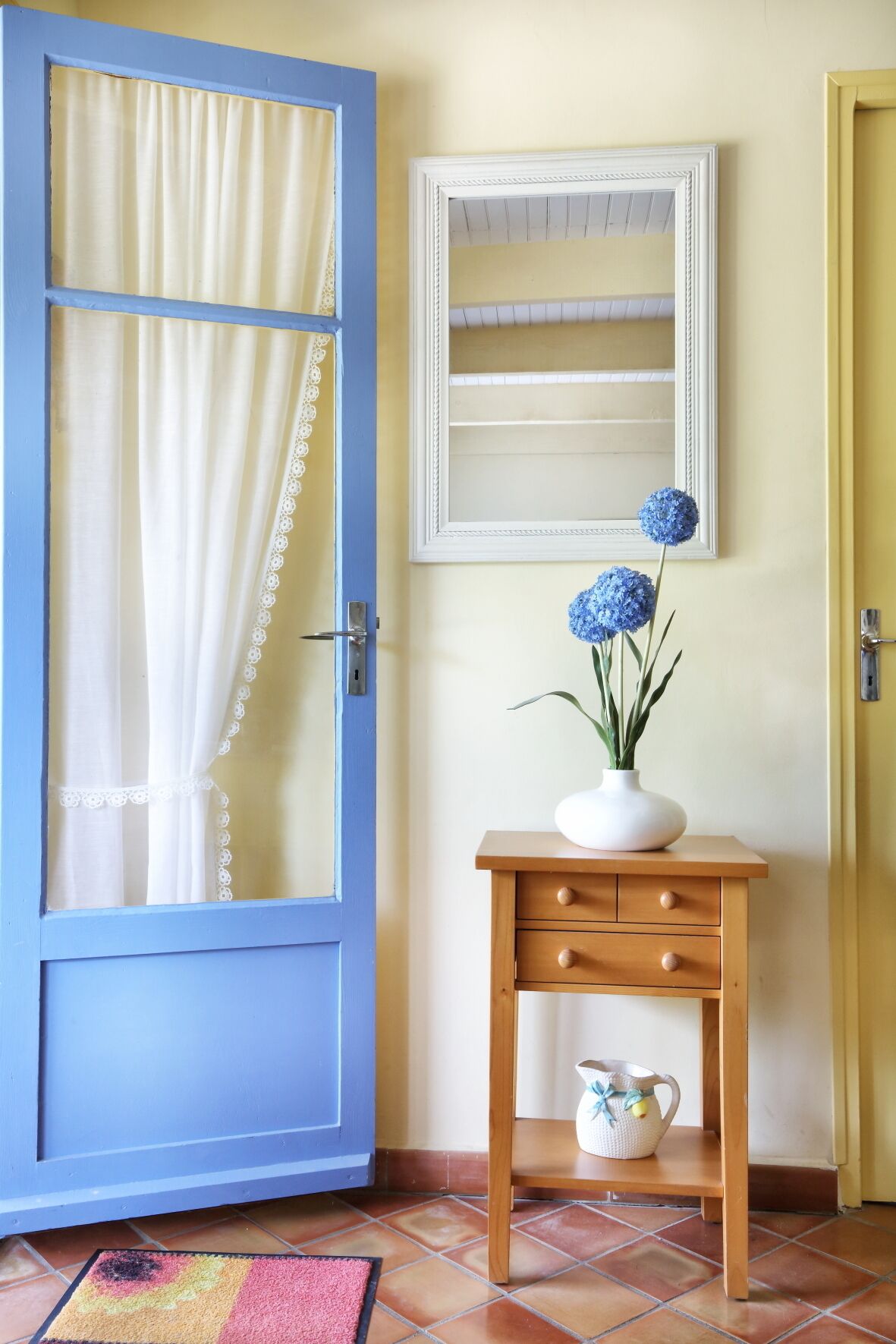 Una puerta azul con una cortina blanca, un jarrón blanco con flores azules, una mesita de madera y un suelo de baldosas.
