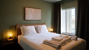 Habitación de hotel con una cama, dos lámparas de noche, un ventanal con cortinas y una obra de arte colgada en la pared.