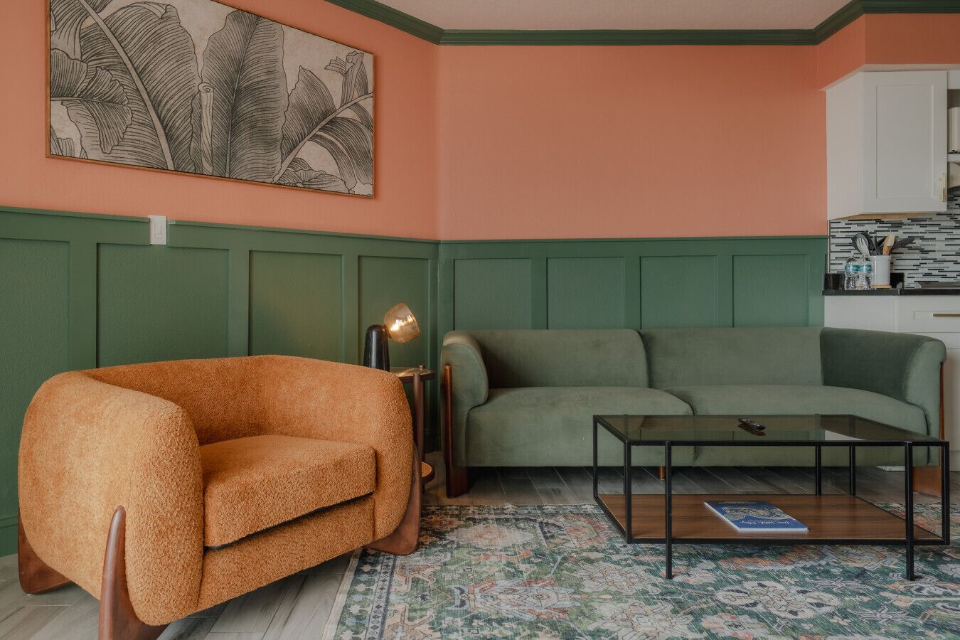 Una sala de estar con un sofá verde, un sillón marrón, una mesa de centro de vidrio y un tapete estampado.