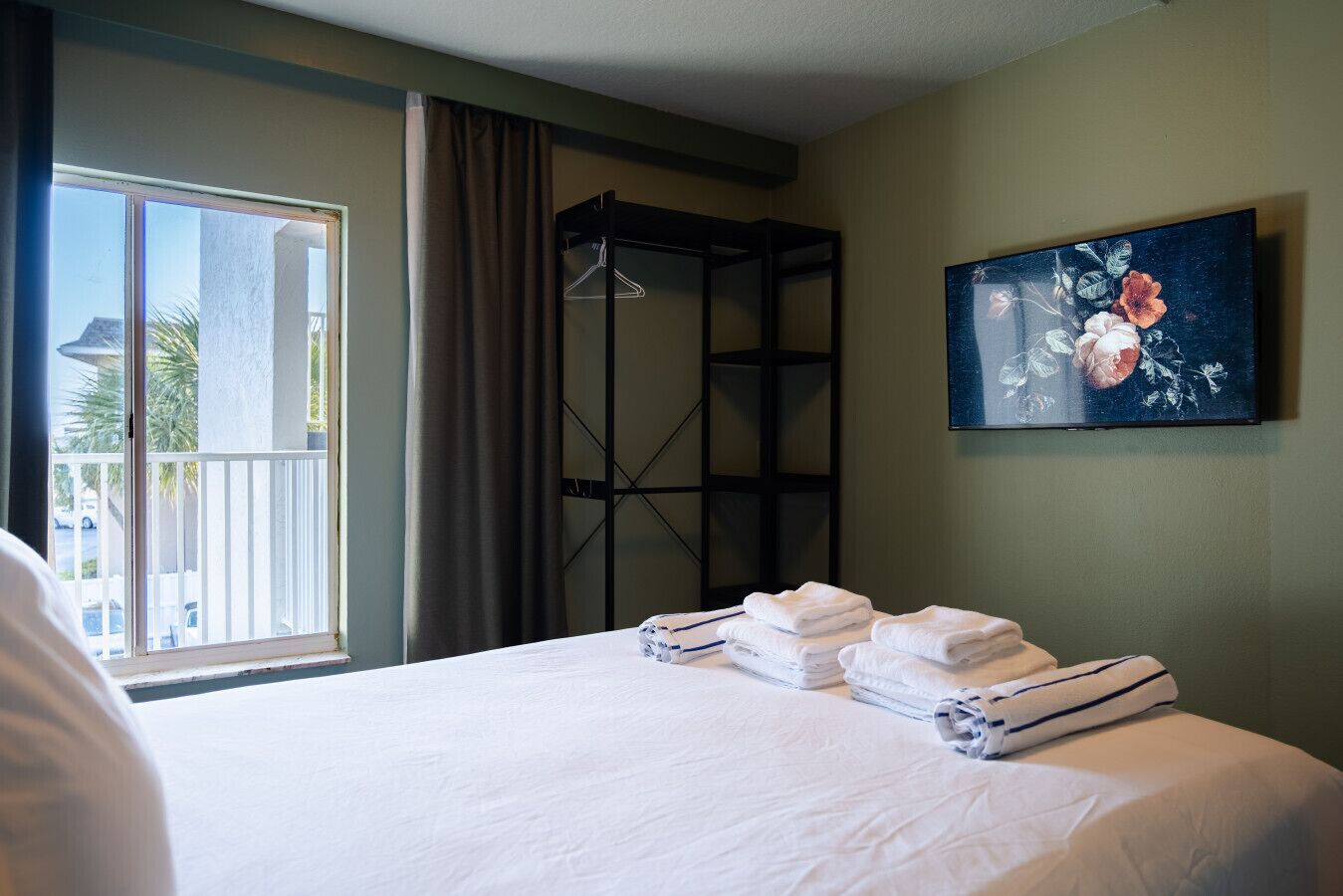 A hotel room with a bed, a window with curtains, a painting on the wall, and a metal rack.