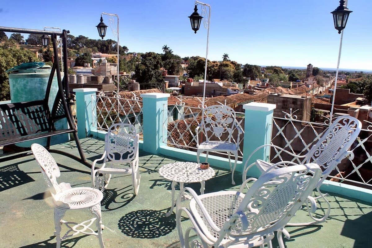 A rooftop terrace with white metal chairs and tables, a metal fence, and a view of a cityscape with buildings and trees.