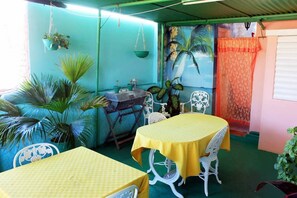 A room with two yellow tables and white chairs, a hanging plant, and a mural of palm trees. - Casa Vista  (Trinidad)