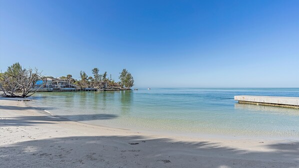 A beach with clear water, a concrete walkway, and buildings in the background.