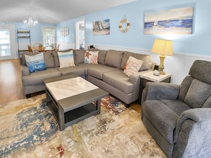 Uma sala de estar com sofá seccional, mesa de centro, tapete estampado e obras de arte na parede, incluindo uma placa com a inscrição 'Beach House'.