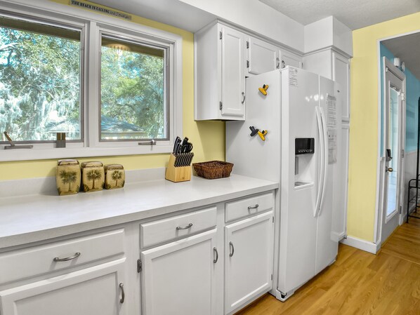 Una cocina con armarios blancos, un ventanal con vistas a árboles y un refrigerador con imanes.