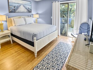 Quarto com cama, duas luminárias de cabeceira, um quadro, varanda com uma cadeira e escrivaninha com computador.