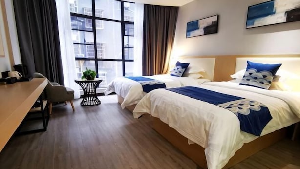 Hotellihuone, jossa on kaksi sänkyä, työpöytä, tuoli, pieni pöytä, jossa on kasvi, ja suuri ikkuna, jossa on verhot.