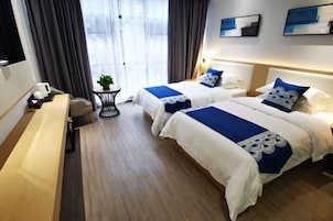 Habitación de hotel con dos camas, una mesita, un banco y vistas a la ciudad a través de amplios ventanales.