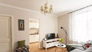 Ein modernes Wohnzimmer mit einer Couch, einem Fernseher auf einem weißen Schrank, einem kleinen Beistelltisch mit Blumen und einer Chandelüster.