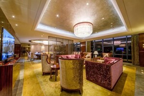 A spacious lobby with a large chandelier, plush seating, a television, and a view of the city at night.