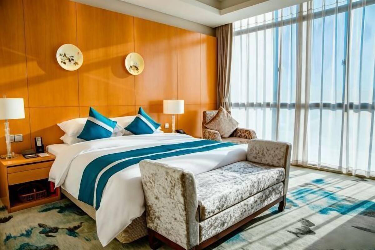 Een hotelkamer met een groot bed, twee nachtkastjes met lampen, een gestreept fauteuil en een groot raam met gordijnen.