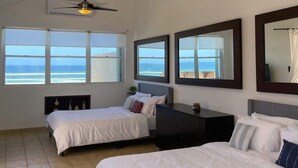A hotel room with two beds, a ceiling fan, a large window with a view of the ocean, and a mirror on the wall.
