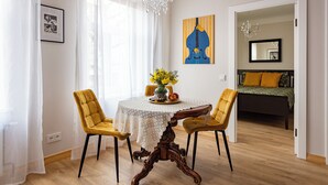 Een eethoek met een ronde tafel, twee gele stoelen, een kroonluchter en een schilderij aan de muur.