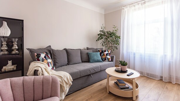 Een moderne woonkamer met een grijze bank, een ronde salontafel, een roze fauteuil en een boekenkast met decoratieve objecten.