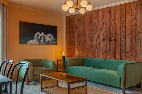 Ein Wohnzimmer mit einem grünen Sofa, einem Glastisch und einem Gemälde von Wolken an der Wand.